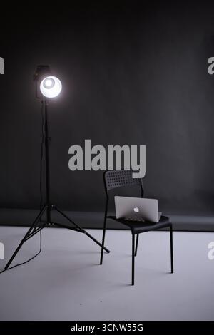 A minimalist studio scene featuring a professional photo light on a tripod and a laptop resting on a black chair against a dark gray background Stock Photo