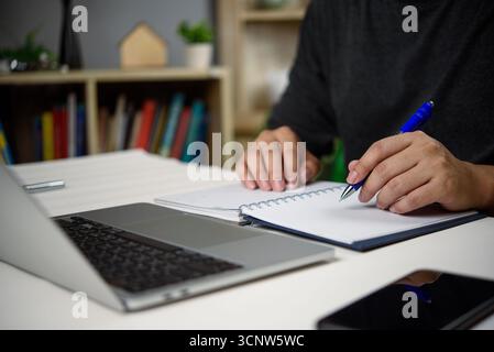 Close-up of hands writing in notebook while working on laptop, and smartphone in a modern home office. planning, and productive workflow. concept for Stock Photo