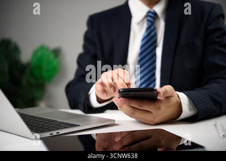 Businessman using smartphone at desk with laptop, mobile communication, business multitasking, and digital productivity in a corporate environment. Stock Photo