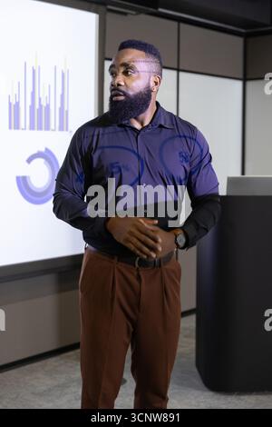 African american man presenting charts on screen in meeting room in formal attire at podium Stock Photo