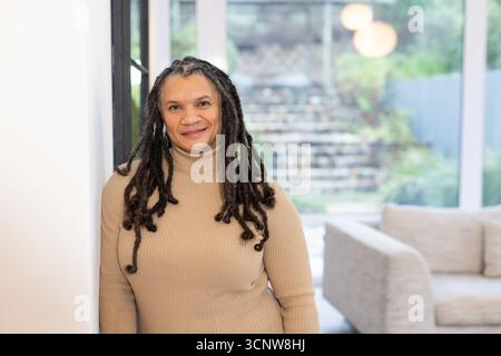 Senior African American woman leaning against white wall in living room near glass door, copy space Stock Photo