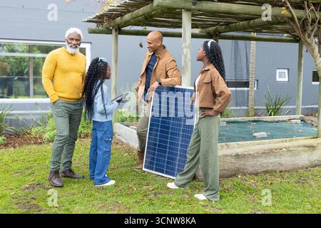 Diverse family gathering around solar panel under pergola on grassy lawn with tablet device Stock Photo