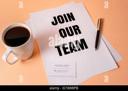White paper with "Join Our Team" text beside coffee mug and business card on desk. Stock Photo