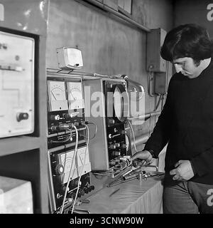 A focused Soviet engineer or technician works intently with a complex electronics test bench in a laboratory in Sloviansk, Ukrainian SSR, during the 1980s. The specialist is adjusting controls on the vintage equipment, which features an oscilloscope, analog dials, and a web of wiring, likely performing diagnostics or calibration. This black-and-white photograph captures a moment of intense concentration, showcasing the skilled, high-tech side of the Soviet industrial and scientific workforce Stock Photo