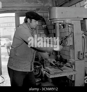 A skilled Soviet machinist concentrates on his work, operating a large, heavy-duty drill press in a machine shop in Sloviansk, Ukrainian SSR, during the 1980s. Dressed in a classic work jacket and cap, the man carefully drills a metal part, while a "Kachestvo" (Quality) sign is visible in the background—a common motivational slogan in USSR factories. This black-and-white photograph authentically captures the atmosphere of a blue-collar profession and the focus on industrial production in the late Soviet era Stock Photo
