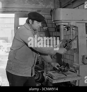 A skilled Soviet machinist concentrates on his work, operating a large, heavy-duty drill press in a machine shop in Sloviansk, Ukrainian SSR, during the 1980s. Dressed in a classic work jacket and cap, the man carefully drills a metal part, while a "Kachestvo" (Quality) sign is visible in the background—a common motivational slogan in USSR factories. This black-and-white photograph authentically captures the atmosphere of a blue-collar profession and the focus on industrial production in the late Soviet era Stock Photo