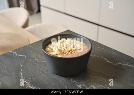 Bright and inviting banana-strawberry smoothie sits in a dark bowl, garnished with fresh banana slices and pine nuts. The cozy kitchen provides a warm Stock Photo
