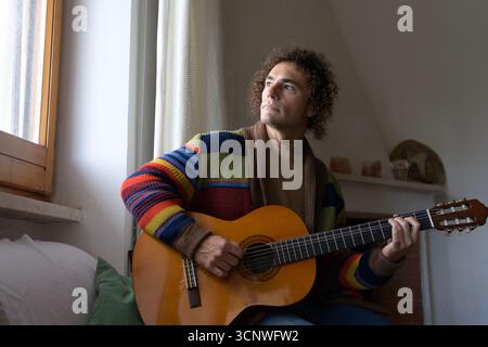 A man wearing a colorful sweater plays an acoustic guitar by the window. Relaxing at home, he strums the strings, immersed in the music, enjoying the Stock Photo