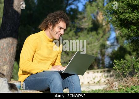 A man in a yellow sweater sits outdoors in a sunny garden, using a laptop. He is focused and smiling, enjoying the warm weather and the fresh air. Stock Photo