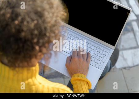 A man in a yellow sweater sits comfortably at home using a white laptop. The focus is on his hands typing on the keyboard, set against a relaxed, resi Stock Photo