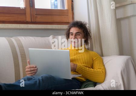A man in a yellow sweater sits comfortably on a couch at home, engaged with his laptop. Natural light filters through a window, creating a cozy and in Stock Photo