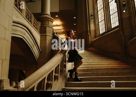 A woman in autumn attire stands thoughtfully on an ornate staircase, illuminated by soft light filtering through large windows, showcasing Seattle's a Stock Photo