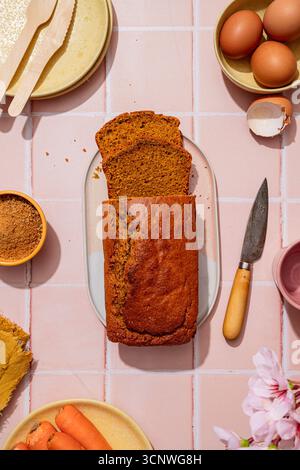 Top view of a homemade carrot cake loaf on a pink tiled background. Surrounded by eggs, a knife, sugar, carrots, and flowers, creating a rustic and in Stock Photo