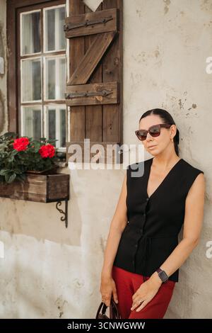 Woman in sunglasses stands leisurely against a rustic wall, beside a wooden shuttered window adorned with vibrant red flowers, embodying casual elegan Stock Photo
