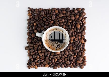 A white coffee cup filled with dark coffee is centered on a square arrangement of roasted coffee beans on a white background, creating a simple and el Stock Photo