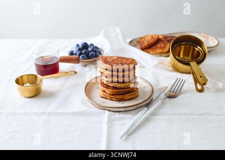 A stack of golden pancakes sits on a plate, surrounded by fresh blueberries, syrup, and brass measuring cups; embodying a serene breakfast atmosphere Stock Photo