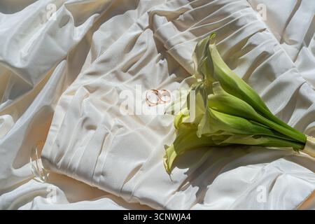 Elegant wedding rings rest on a background of luxurious white silk fabric, accompanied by a fresh green bouquet. The setting embodies a sense of roman Stock Photo