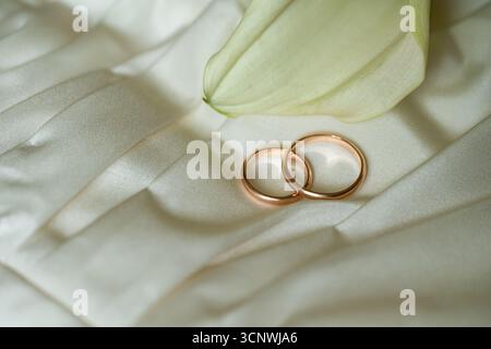 Two elegant gold wedding rings rest delicately on a smooth cream satin fabric, accompanied by a pale green calla lily flower, symbolizing love and com Stock Photo