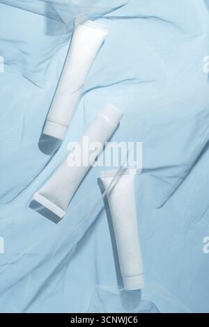 Top view of three white cosmetic cream tubes elegantly placed on a light blue background, covered by sheer mesh fabric, showcasing the minimal design Stock Photo