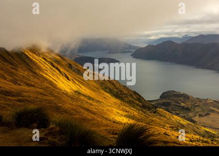 Golden sunlight breaks through clouds over the slopes of Roys Peak in Wanaka, New Zealand, during autumn. The dramatic view of Lake Wanaka combines mi Stock Photo