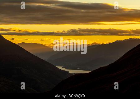 Fiery orange sunset colors fill the autumn sky over Lake Wakatipu in Queenstown, New Zealand. Layers of shadowed mountains add depth and mood to this Stock Photo