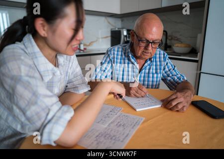 A senior man engaged in learning Mandarin, guided by an asian woman teacher, in a cozy kitchen setting. Both are focused on language notes and study m Stock Photo
