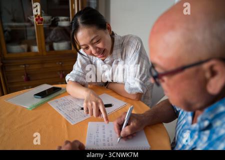 A senior man learns Mandarin with the help of an Asian woman teacher. They are seated at a table, engaged in a one-on-one learning session with notes Stock Photo