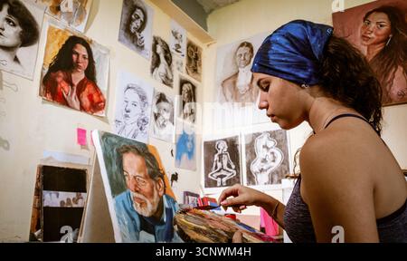 A young female artist focuses on her painting in a personal studio, surrounded by expressive artwork. The atmosphere is filled with creativity and ins Stock Photo
