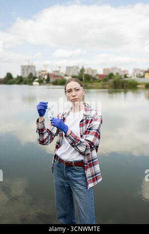 A young female biologist wearing safety glasses and gloves conducts field research by a scenic lake. She holds a test tube, gathering water samples fo Stock Photo