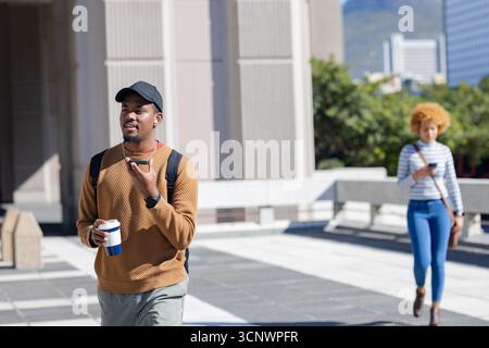 African american man striding across plaza holding reusable coffee cup, smartphone, copy space Stock Photo