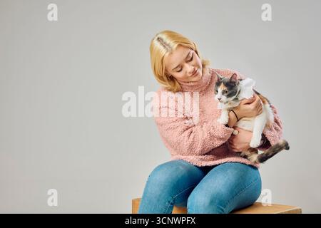 A blonde woman in a cozy pink sweater sits on a wooden box in a studio, lovingly holding a cat dressed in a cute accessory, conveying warmth and compa Stock Photo