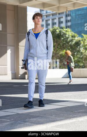 Man standing under plaza canopy wearing tracksuit and holding skateboard, backpack and earbuds Stock Photo