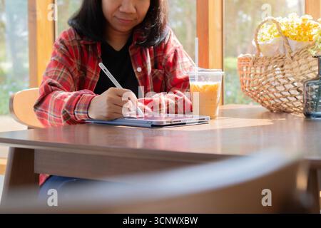 Adult Asian woman sitting in coffee shop at wooden table, drinking coffee and using tablet. Lifestyle. Technology concept Stock Photo