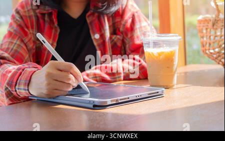 Close-up woman sitting on the wooden table with iced coffee and using digital tablet in a cafe. Selective focus. Stock Photo