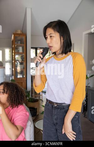 Asian mother singing into wireless microphone at home while African American daughter sipping drink Stock Photo