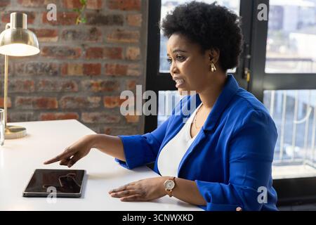 African american woman in blue blazer leaning, pointing at tablet on white desk in office Stock Photo