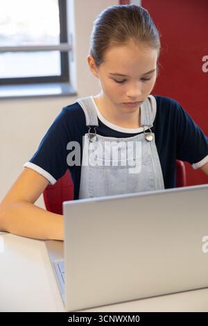 Teenage female student studying on laptop at white desk with red chair inside classroom Stock Photo