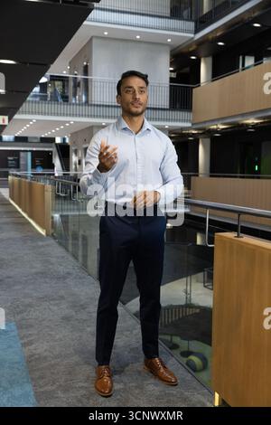 Indian man standing next to glass railing in corporate atrium, gesturing amid ceiling lights Stock Photo