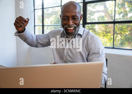 Senior african american man celebrating success at desk with laptop under window in office Stock Photo