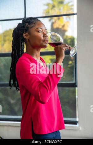 African american woman wearing pink blouse by grid window holding red wine glass Stock Photo