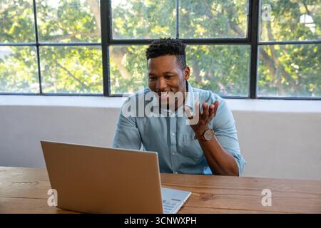 African american man in shirt using laptop at wooden table by window with green foliage Stock Photo