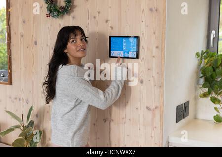Hispanic woman adjusting touch panel on wood wall with wreath, window, plant, outlets Stock Photo