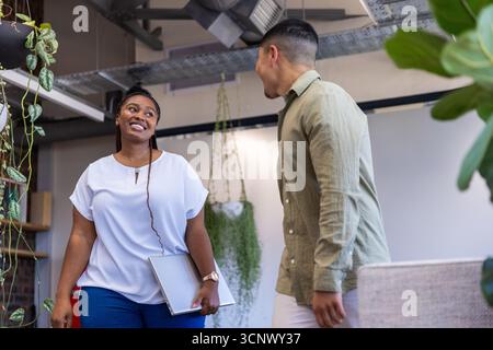 Diverse coworkers smiling and holding closed laptop in office with planters Stock Photo