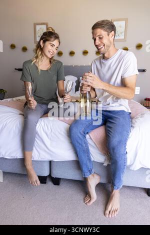 Couple sitting on bed in room holding champagne bottle and empty flutes beneath botanical prints Stock Photo