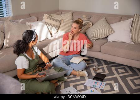Diverse female friends sitting on rug in living room corner collaborating with laptop, charts Stock Photo