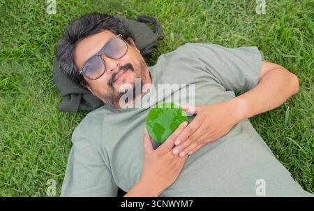 Top view of happy handsome beard man wearing eye grasses holding a green earth ball while lying on the lawn. World environment day and global conserva Stock Photo