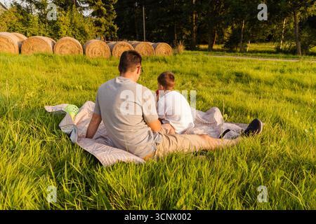 A father and son sit together on a blanket in a grassy field, enjoying quality time in nature. They appear relaxed as they share stories and laughter Stock Photo