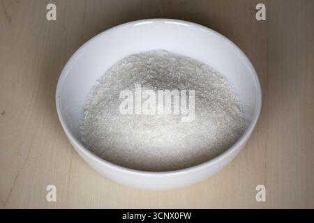 A white bowl is filled with sugar on a wooden textured background. Stock Photo