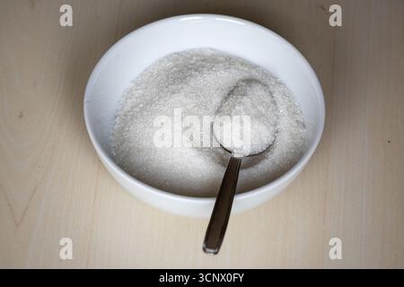 A white ceramic bowl and steel spoon full of sugar on a wooden background. Ingredients used in cooking sweet foods. Stock Photo