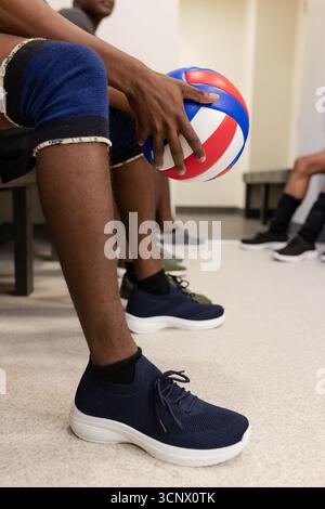 Diverse male volleyball team sitting in locker room holding volleyball in knee pads, sneakers Stock Photo